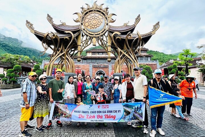 Một nhóm đông người đứng chụp ảnh tại cổng vào với kiến trúc độc đáo, có mái vòm lớn và hoa văn tinh xảo. Trời mát mẻ, phía sau là đồi núi xanh tươi. Mọi người đều vui vẻ, cầm biển hiệu "Du Lịch Đà Nẵng" với ngày tháng rõ ràng. Nhóm du khách chụp ảnh tại Bà Nà Hills, Đà Nẵng, với biển hiệu "Du Lịch Đà Nẵng".