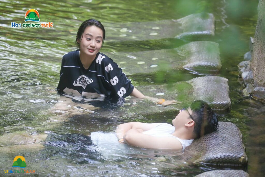 Cặp đôi trò chuyện và thư giãn trong dòng suối nước nóng, tận hưởng không gian yên tĩnh của công viên suối khoáng nóng Núi Thần Tài.