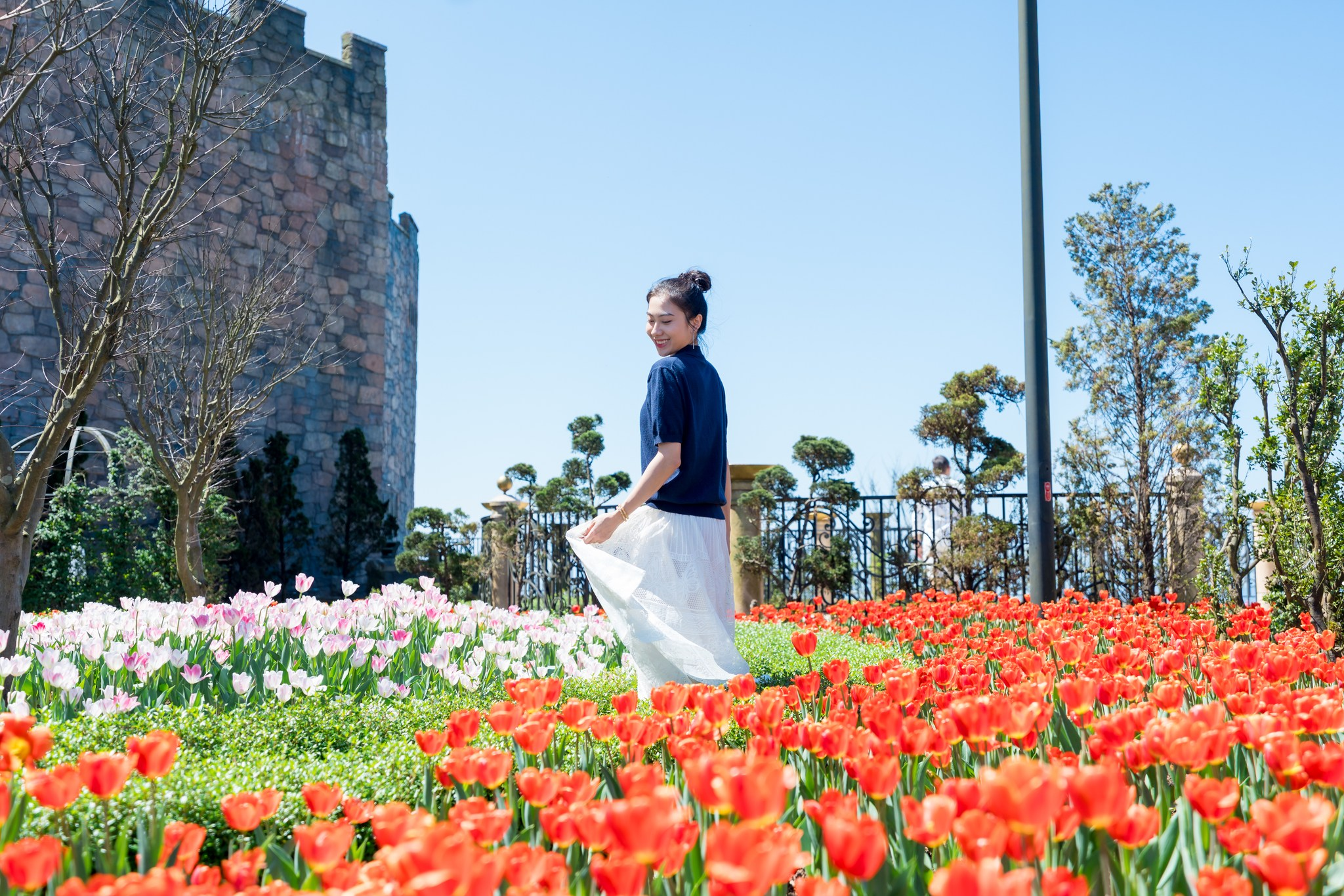 Cô gái đứng giữa một vườn hoa tulip rực rỡ với nhiều màu sắc khác nhau, chủ yếu là đỏ và trắng. Ánh nắng chiếu sáng làm nổi bật vẻ tươi tắn của hoa. Cô mặc áo tối màu, váy trắng, đang mỉm cười và quay lại về phía camera, tạo dáng tự nhiên. Phía sau là một bức tường đá cổ kính và cây cối xanh um, tạo nên không gian lãng mạn. Cô gái tạo dáng giữa vườn hoa tulip rực rỡ tại Bà Nà Hills.