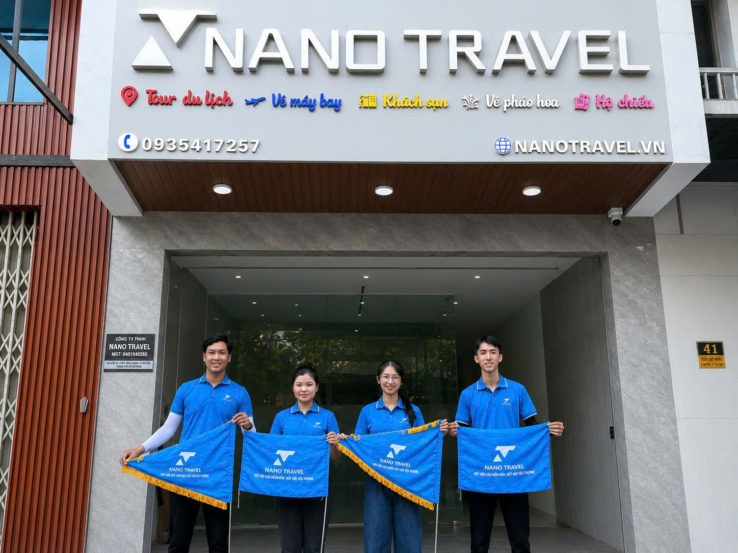Đội ngũ nhân viên của Nano Travel đứng trước văn phòng, cầm cờ hiệu với logo công ty, thể hiện sự chuyên nghiệp và sẵn sàng phục vụ khách hàng.
