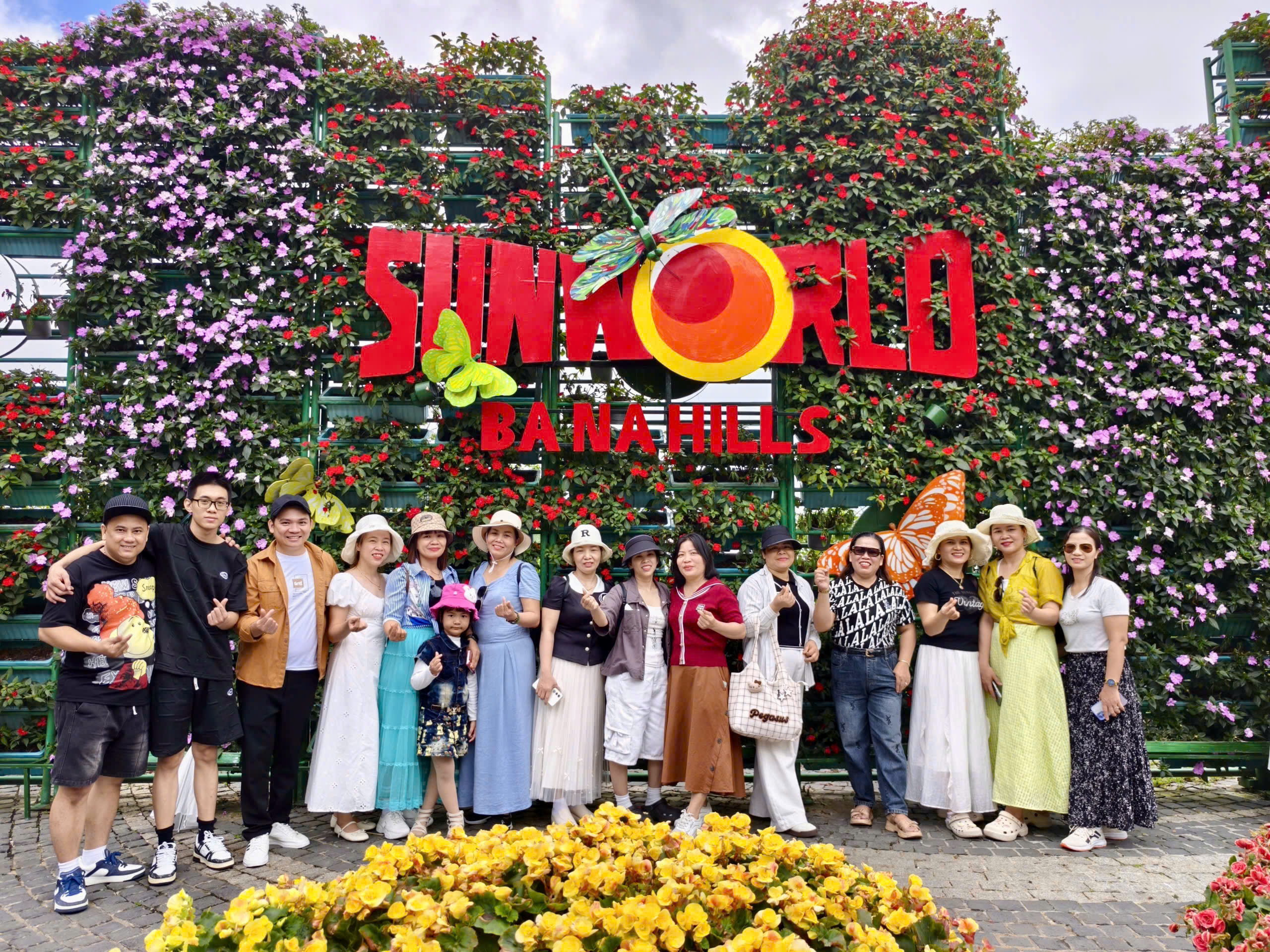 Nhóm khách du lịch chụp ảnh kỷ niệm tại cổng vào Sun World Ba Na Hills, với nhiều hoa và cây xanh rực rỡ xung quanh, thể hiện năng lực tổ chức đoàn lớn của vebanahill.vn trong hành trình 400 khách Yuri ABC.